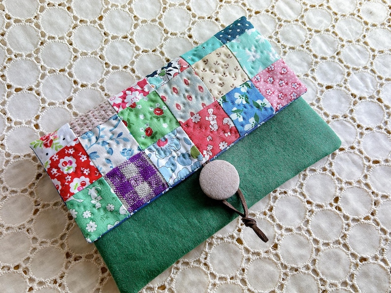 Pode incluir: Uma clutch de tecido patchwork com base em feltro verde. O patchwork apresenta v&aacute;rios tecidos florais e estampados em vermelho, azul, verde e rosa. Um bot&atilde;o bege e um fecho de cord&atilde;o castanho s&atilde;o vis&iacute;veis.