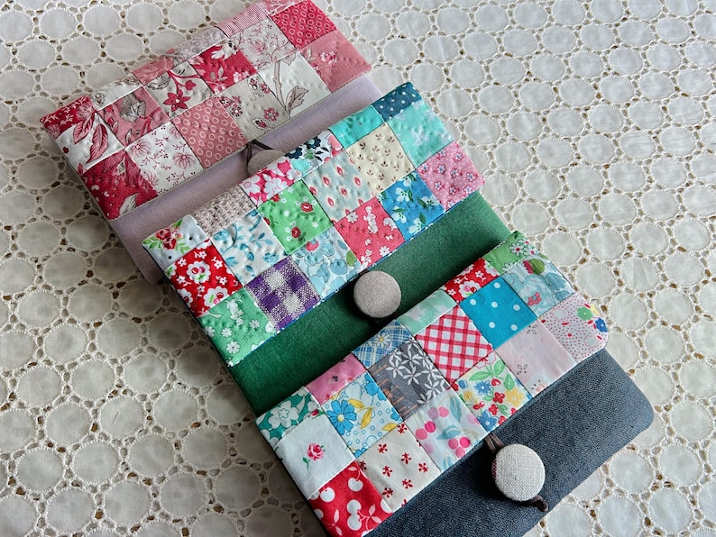 Pode incluir: Estojos de tecido patchwork feitos &agrave; m&atilde;o em v&aacute;rios tamanhos. Os estojos apresentam uma variedade de tecidos florais e estampados coloridos, com fecho de bot&atilde;o. Os estojos est&atilde;o dispostos sobre uma toalha de mesa de renda branca.