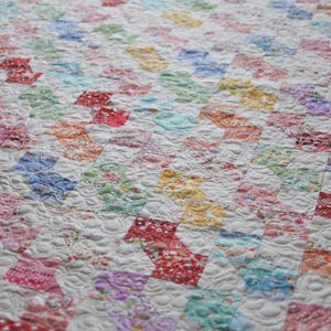Könnte beinhalten: Ein Steppdecke mit einem sich wiederholenden Muster aus kleinen, bunten Stoffquadraten in Rot-, Blau-, Gelb-, Grün- und Rosatönen, durchsetzt mit weißen Quadraten. Die gesamte Oberfläche ist mit einer dichten, wirbelnden Steppnaht versehen.