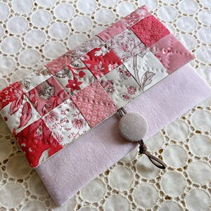 Pode incluir: Uma clutch acolchoada feita &agrave; m&atilde;o com um design de patchwork em tons de vermelho, rosa e branco com padr&otilde;es florais. A clutch tem uma base de feltro roxo claro e um fecho de bot&atilde;o com um cord&atilde;o castanho. O fundo &eacute; uma toalha de mesa de renda branca.