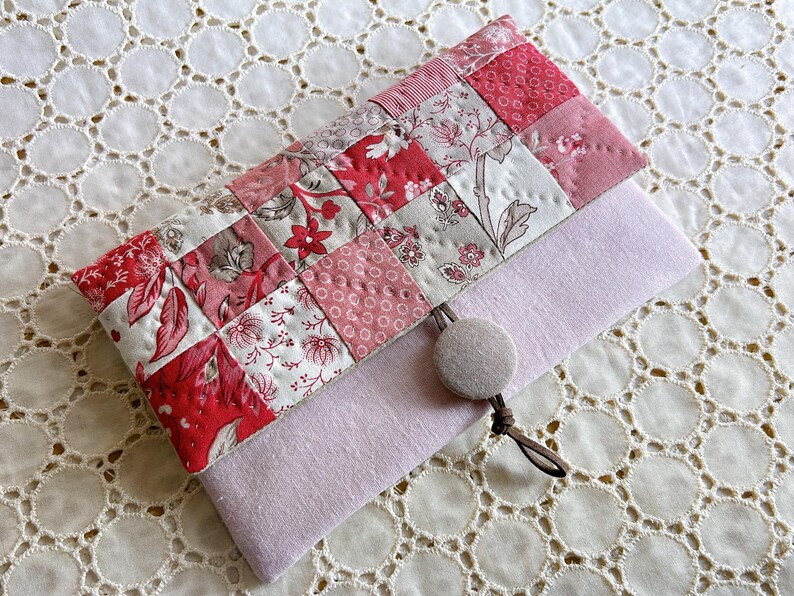 Pode incluir: Uma clutch acolchoada feita &agrave; m&atilde;o com um design de patchwork em tons de vermelho, rosa e branco com padr&otilde;es florais. A clutch tem uma base de feltro roxo claro e um fecho de bot&atilde;o com um cord&atilde;o castanho. O fundo &eacute; uma toalha de mesa de renda branca.