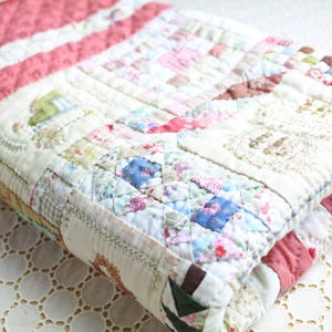 May include: Folded patchwork quilt featuring a variety of floral and geometric patterns in pastel colors. The quilt has a white background with pink and red accents. The edges are finished with a decorative stitch.