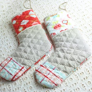 Peut inclure: Deux bas de Noël matelassés. Le corps principal est gris avec un motif en losange. Le bord supérieur est rouge avec des motifs de vacances blancs et bleus. Le bout est un motif à carreaux rouge, blanc et bleu clair. Chaque bas a une boucle pour accrocher.