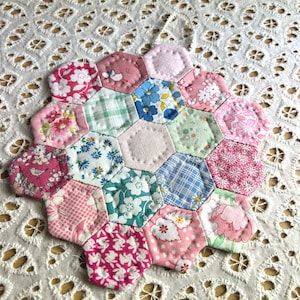 May include: A handmade patchwork coaster featuring a collection of hexagonal fabric pieces. The coaster showcases a variety of floral and patterned designs in shades of pink, blue, and green. It rests on a white, lace-like surface.