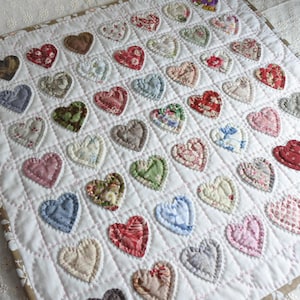 Op de afbeelding: Een handgemaakte quilt met een raster van stoffen harten in verschillende kleuren en patronen, waaronder bloemen- en effen ontwerpen. De harten zijn op een witte achtergrond geappliqueerd met gewatteerde stiksels en de quilt heeft een bruine rand.
