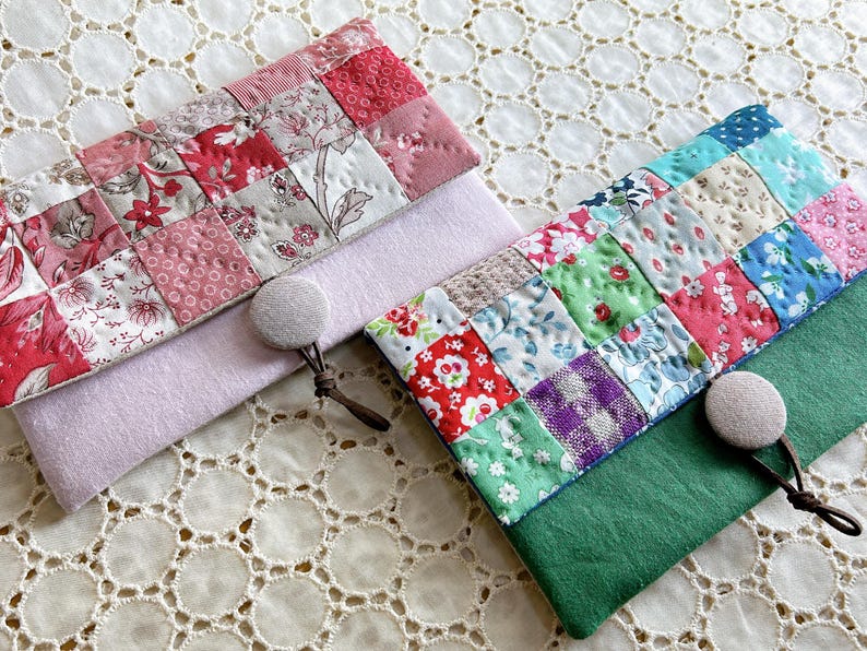 Pode incluir: Duas bolsas de tecido feitas &agrave; m&atilde;o com designs de patchwork. Uma &eacute; rosa e vermelha, a outra &eacute; verde e multicolorida. Cada bolsa tem um fecho de bot&atilde;o e um pequeno la&ccedil;o. As bolsas est&atilde;o sobre uma superf&iacute;cie de renda branca.