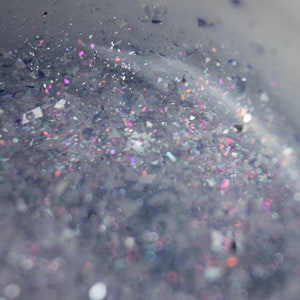 May include: Close-up of a clear liquid filled with iridescent glitter and small, colourful particles. The glitter reflects pink, purple, and silver hues, creating a shimmering effect. The image has a soft, blurred background.