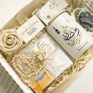 May include: A gift box filled with items for Ramadan. The box contains a white mug with gold Arabic calligraphy, a small box of chocolates, a string of pearls, and a card that says "Ramadan Kareem".