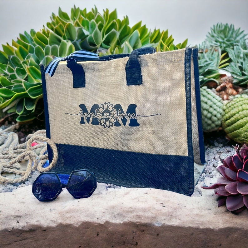 K&ouml;nnte beinhalten: Eine beige Tragetasche mit blauen Einfassungen und Griffen. Die Tasche hat ein schwarz-wei&szlig;es Design mit dem Wort "MOM" und einer Sonnenblumengrafik. Die Tasche liegt auf einer Oberfl&auml;che mit gr&uuml;nen Pflanzen im Hintergrund.