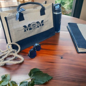 K&ouml;nnte beinhalten: Eine Tragetasche aus Naturfaser mit schwarzen Henkeln und einem schwarzen Boden. Die Tasche hat ein schwarzes gesticktes Design mit der Aufschrift "Mom" und einer Sonnenblumengrafik. Die Tasche liegt auf einem Holztisch mit einer Wasserflasche, einer Sonnenbrille und einem schwarz-wei&szlig; gestreiften Stoff.