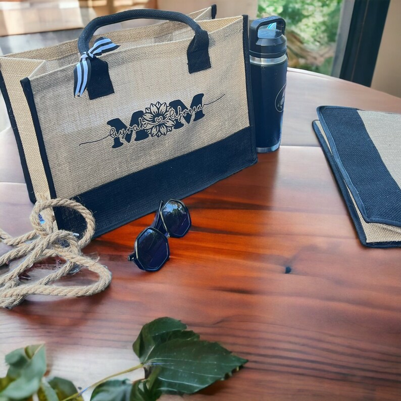 K&ouml;nnte beinhalten: Eine Tragetasche aus Naturfaser mit schwarzen Henkeln und einem schwarzen Boden. Die Tasche hat ein schwarzes gesticktes Design mit der Aufschrift "Mom" und einer Sonnenblumengrafik. Die Tasche liegt auf einem Holztisch mit einer Wasserflasche, einer Sonnenbrille und einem schwarz-wei&szlig; gestreiften Stoff.