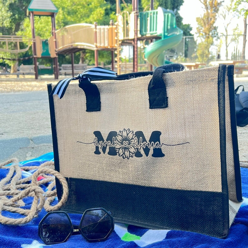 K&ouml;nnte beinhalten: Eine beige und schwarze Tragetasche mit einem schwarz-wei&szlig; gestreiften Griff. Die Tasche hat ein schwarzes Design mit der Aufschrift "Mom" und einer Sonnenblume in der Mitte. Die Tasche liegt auf einem blau-gr&uuml;nen Handtuch.