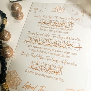 May include: A white card with gold lettering and decorative borders. The card features three different Islamic prayers for the three Ashras of Ramadan. The text includes the Arabic script and English translations.