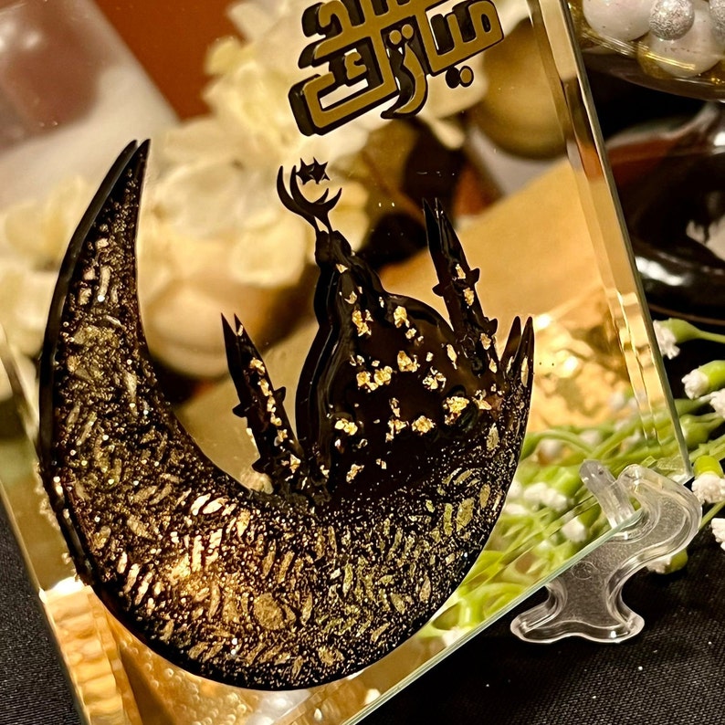 May include: A gold and black glass picture frame with a crescent moon and mosque design. The frame has the Arabic word "Eid" written on it.