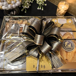 May include: A gold gift basket with a black and gold bow. The basket contains a variety of treats, including nuts, chocolate, and pastries. The basket is wrapped in clear cellophane and tied with a black ribbon. The basket is decorated with a gold card that says "Eid Mubarak".