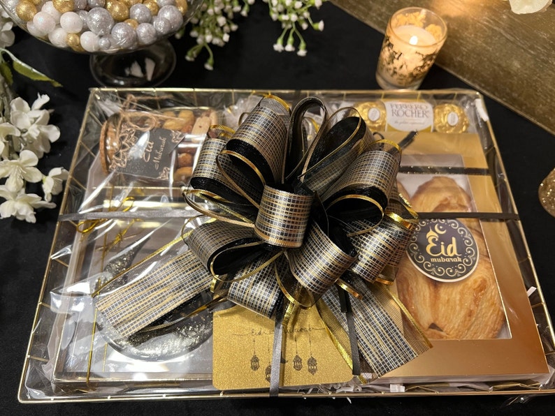 May include: A gold gift basket with a black and gold bow. The basket contains a variety of treats, including nuts, chocolate, and pastries. The basket is wrapped in clear cellophane and tied with a black ribbon. The basket is decorated with a gold card that says "Eid Mubarak".