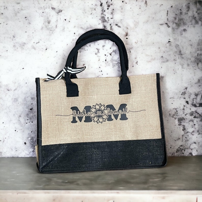 K&ouml;nnte beinhalten: Eine braune und schwarze Jute-Tragetasche mit schwarzen Henkeln. Die Tasche hat einen schwarzen Boden und ein wei&szlig;-schwarzes Band, das an den Henkeln befestigt ist. Die Tasche tr&auml;gt den Text "Mom" in Schwarz mit einer Sonnenblumengrafik und den Text "Bisho & Ismail" in Kursivschrift.