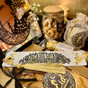 May include: A gold gift box with a black ribbon and a gold bookmark with Arabic calligraphy. The box contains pastries with a gold sticker that says "Eid Mubarak". There is a glass jar filled with mixed nuts and a small bag with a candle.
