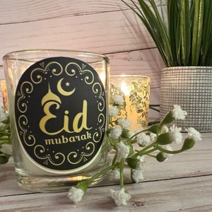 May include: A clear glass candle jar with a black and gold sticker that says "Eid Mubarak". The sticker features a crescent moon and a star.
