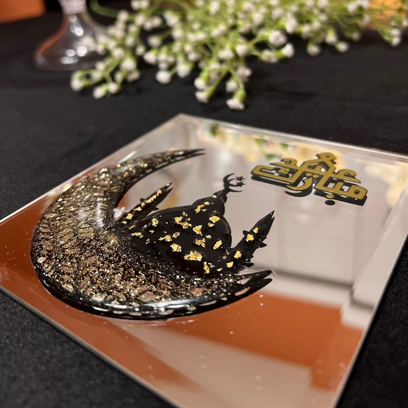 May include: A gold and black resin art piece depicting a crescent moon with a mosque inside. The Arabic text "Eid Mubarak" is written in gold on the right side of the image.