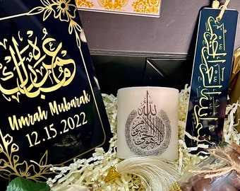 May include: A gift box filled with Islamic-themed gifts, including a black plaque with gold Arabic calligraphy and the text "Umrah Mubarak 12.15.2022", a white candle with gold Arabic calligraphy, a black bookmark with gold Arabic calligraphy, and a glass jar filled with chocolate candies.