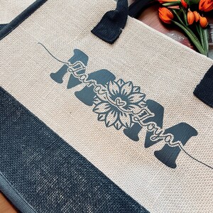 K&ouml;nnte beinhalten: Eine Tragetasche aus Naturfaser mit schwarzen Henkeln und einem schwarzen Design, das "Mama" mit einer stilisierten Sonnenblume in der Mitte sagt.