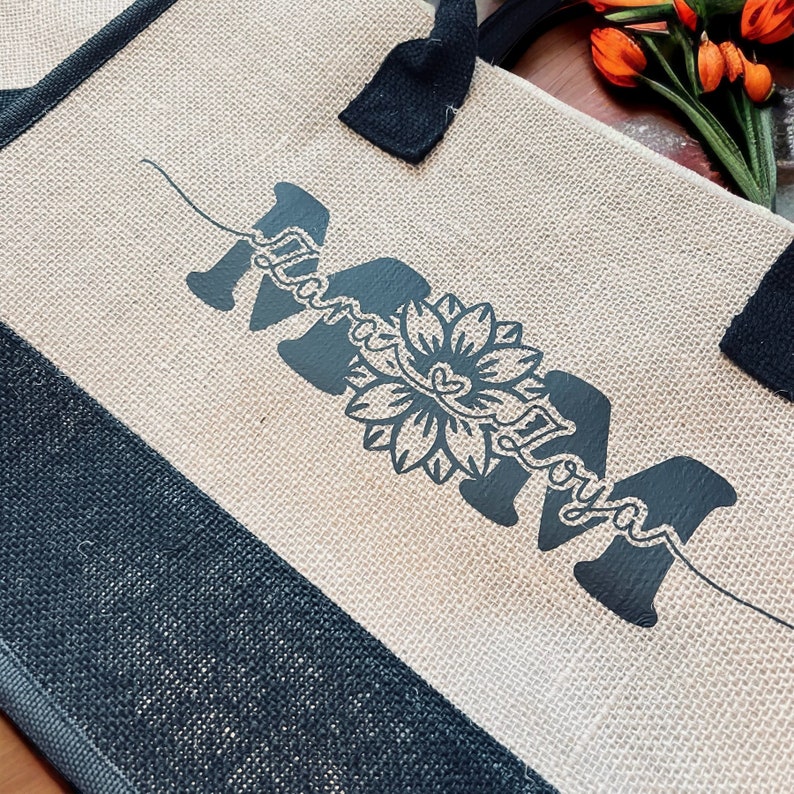 K&ouml;nnte beinhalten: Eine Tragetasche aus Naturfaser mit schwarzen Henkeln und einem schwarzen Design, das "Mama" mit einer stilisierten Sonnenblume in der Mitte sagt.