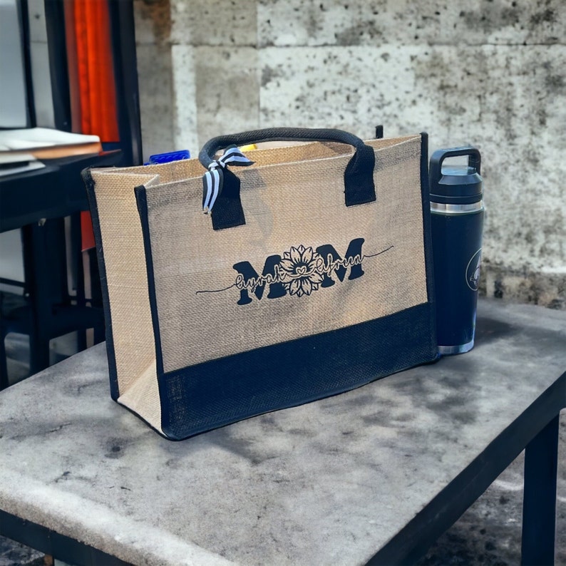 K&ouml;nnte beinhalten: Eine braune und schwarze Tragetasche mit schwarzem Boden und einem Sonnenblumenmuster. Die Tasche tr&auml;gt den Text "Mom" mit einem Sonnenblumenmuster und die Worte "every little thing" auf beiden Seiten der Sonnenblume.