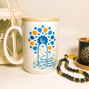 May include: White ceramic mug with a wooden lid. The mug features a blue and orange geometric design with Arabic calligraphy that reads "Ramadan Kareem".