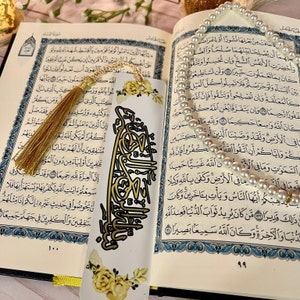 May include: A white bookmark with gold calligraphy and yellow floral accents. The bookmark has a gold tassel attached to the top. The bookmark is laying on top of an open book with Arabic text.