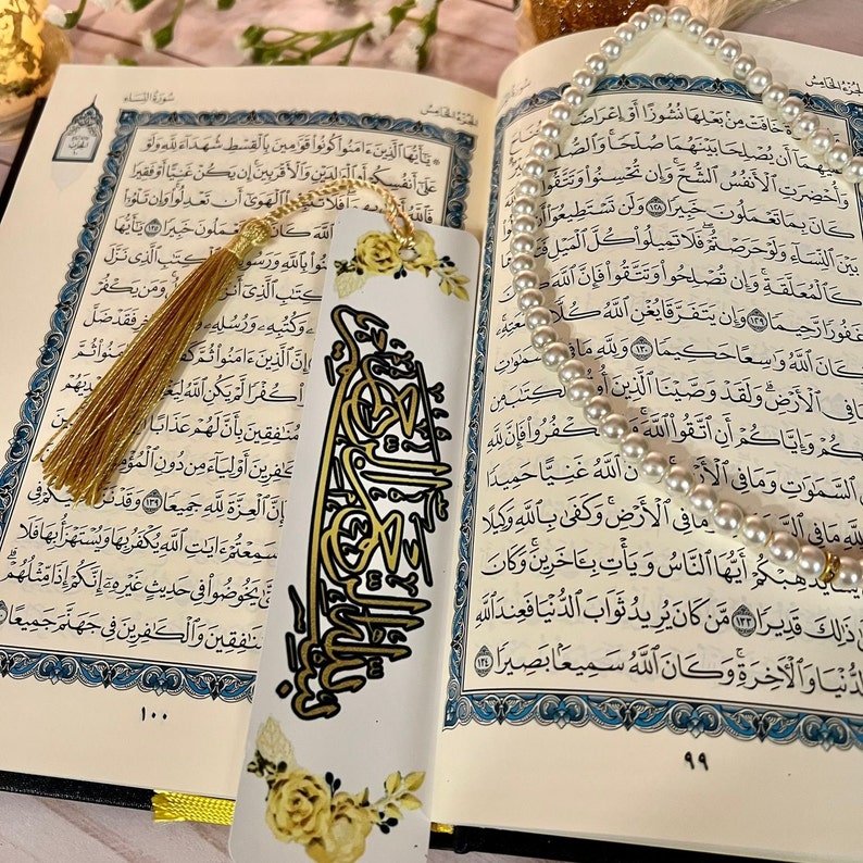 May include: A white bookmark with gold calligraphy and yellow floral accents. The bookmark has a gold tassel attached to the top. The bookmark is laying on top of an open book with Arabic text.