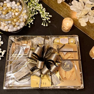 May include: A gold gift basket filled with various treats, including a box of chocolates, a pastry, and a jar of nuts. The basket is decorated with a black and gold bow and a tag that says "Eid Mubarak".