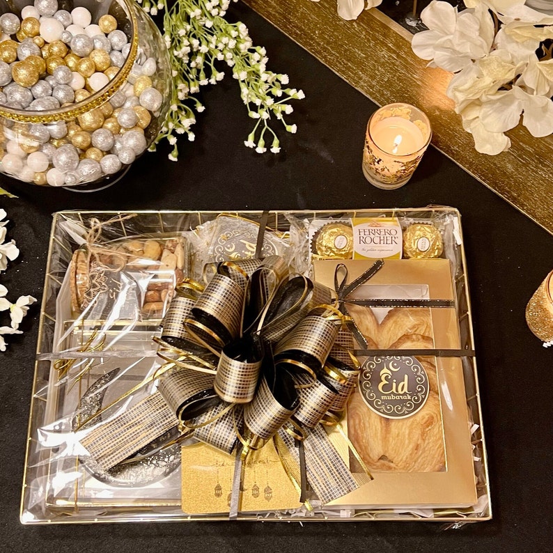 May include: A gold gift basket filled with various treats, including a box of chocolates, a pastry, and a jar of nuts. The basket is decorated with a black and gold bow and a tag that says "Eid Mubarak".