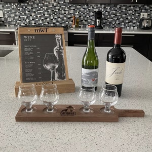 May include: A wooden wine tasting tray with four clear glass tasting glasses and a label that reads "Cattedrale, Special Anniversary". Two bottles of wine are visible in the background, one with a white label that reads "Josh" and the other with a green label.