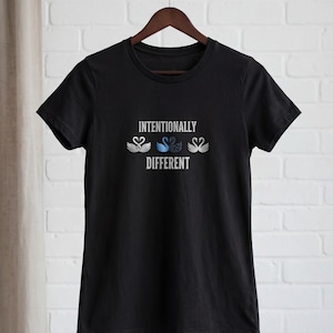 May include: Black t-shirt with the text "Intentionally Different" and three swan graphics in white and blue. The t-shirt is hanging on a wooden hanger against a white brick wall.