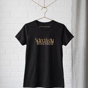 May include: Black t-shirt with the gold text "Selectively Invested" in a stylish font. The shirt is hanging on a metal hanger against a white brick wall. The t-shirt is a classic crew neck style.