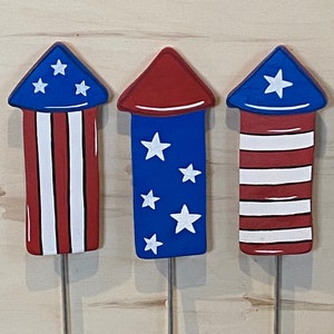 May include: Three wooden firework decorations painted in red, white, and blue with stars. The decorations are attached to metal skewers.