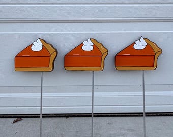 May include: Three wooden garden stakes shaped like pumpkin pie slices. Each slice has an orange filling, a brown crust, and a dollop of white whipped cream. The stakes are made of metal.