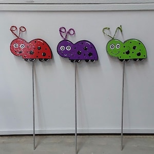 May include: Three colorful ladybug garden stakes. The ladybugs are red, purple, and green with black spots. Each ladybug has a smiling face and is mounted on a metal stake. The ladybugs have glittery antennae.