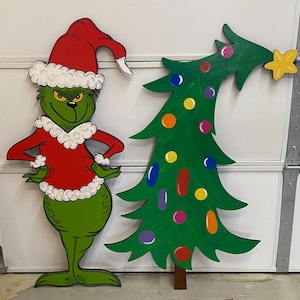 May include: A green Grinch wearing a red Santa suit and a white hat stands next to a green Christmas tree with colorful ornaments.