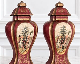 Peut inclure: Une paire de vases couverts assortis avec une finition rouge profond et des bords dorés vieillis. Chaque vase présente un panneau ovale central orné d'un paysage peint avec des arbres et du feuillage, rehaussé de volutes dorées.