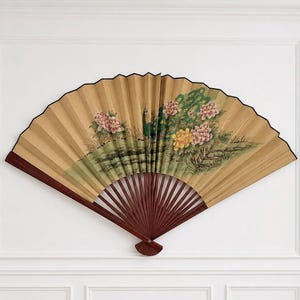 May include: A large, open folding fan featuring a tan paper surface with a painted scene of a green peacock standing among pink and yellow peony flowers. The fan has dark brown wooden ribs and a black border along the top edge of the paper.