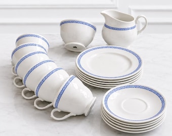 Peut inclure: Un service à thé en porcelaine blanche orné d'un motif grec bleu sur les bords. L'ensemble comprend six tasses avec anses, six soucoupes assorties, un petit pot à lait avec anse et un petit bol, disposés sur une surface blanche.