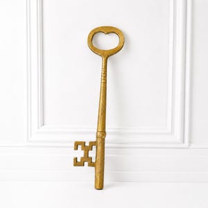 May include: A large, decorative skeleton key finished in a metallic gold tone. The key features a rounded bow at the top, a long, tapered cylindrical shaft with subtle horizontal detailing, and a complex, geometric bit at the bottom end.