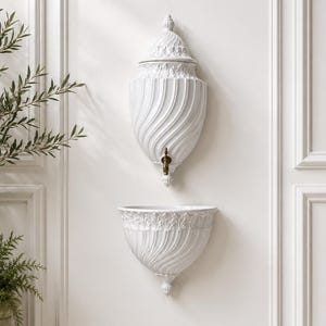 May include: A white wall-mounted water fountain featuring a vertical urn-shaped reservoir with a lid and a matching basin below. The ceramic pieces display a swirling fluted pattern and ornate leaf detailing. A small brass spigot is attached.