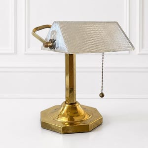 May include: A brass desk lamp featuring an octagonal base, a hexagonal column, and a rectangular, horizontally ribbed glass shade. The lamp includes a brass pull chain with a small spherical end and a side-mounted adjustment knob on the shade.
