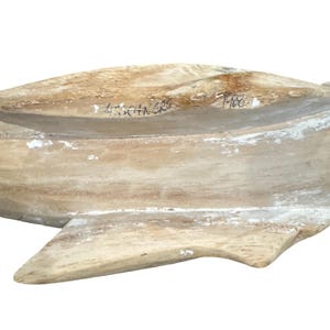 May include: A carved wooden sculpture of a fish with a weathered, light brown and white finish. The surface displays visible wood grain and texture. Handwritten black ink markings are present on the top section, including the year 1988.