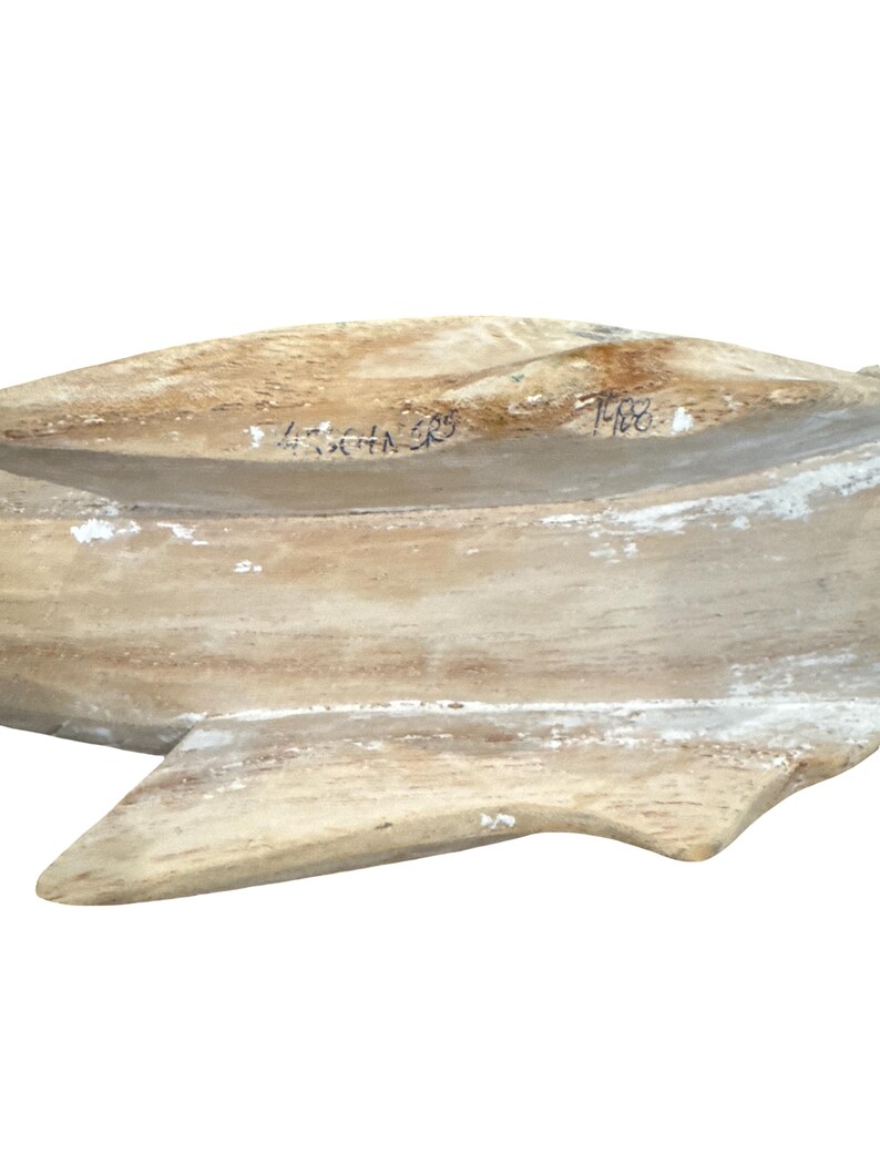 May include: A carved wooden sculpture of a fish with a weathered, light brown and white finish. The surface displays visible wood grain and texture. Handwritten black ink markings are present on the top section, including the year 1988.