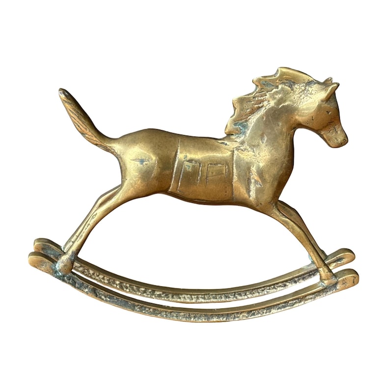 Puede incluir: Una figura de caballo balanc&iacute;n de lat&oacute;n con acabado dorado met&aacute;lico y p&aacute;tina visible. El caballo aparece en posici&oacute;n de galope con crines texturizadas, detalle de silla de montar en el lomo y balancines curvos con un patr&oacute;n decorativo.