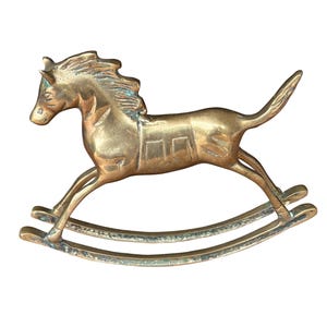 Puede incluir: Una figura de caballo balanc&iacute;n de lat&oacute;n con un acabado met&aacute;lico pulido y p&aacute;tina visible en las hendiduras. El caballo est&aacute; representado en una pose de galope con crines y cola fluidas, montado sobre dos balancines curvos.
